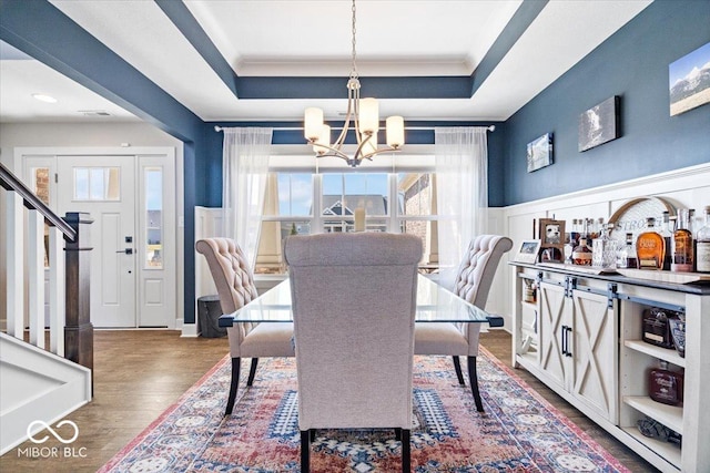 dining area with a tray ceiling, dark wood-type flooring, an inviting chandelier, and wainscoting