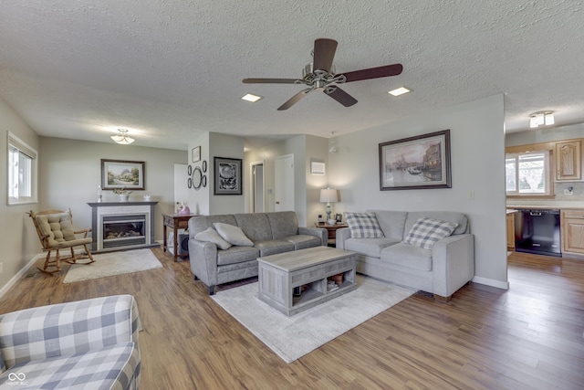 living area featuring baseboards, light wood-style flooring, ceiling fan, a textured ceiling, and a glass covered fireplace