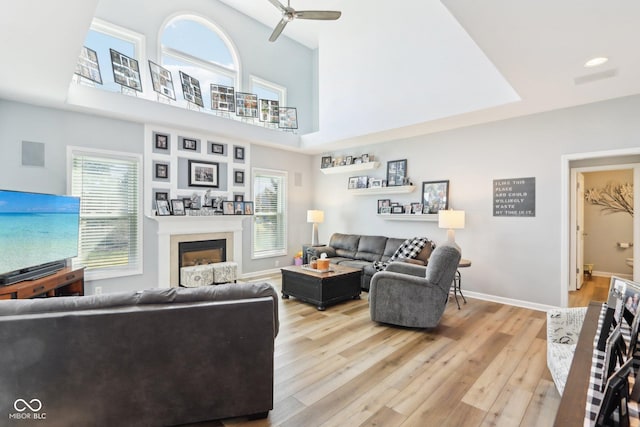 living area featuring a ceiling fan, baseboards, light wood-style flooring, a fireplace, and a towering ceiling