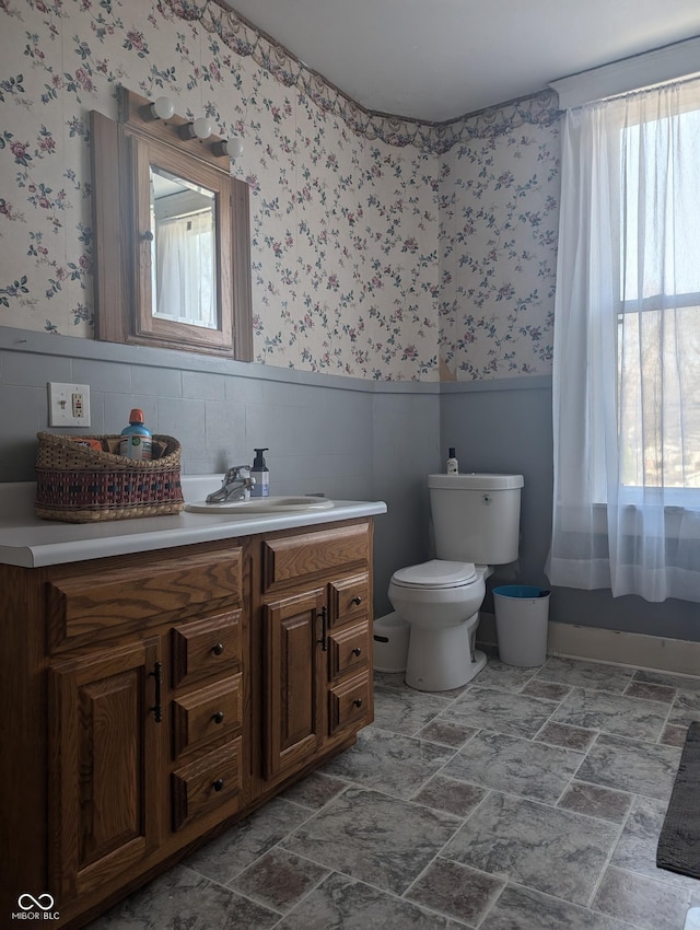 bathroom featuring wallpapered walls, stone finish flooring, a wainscoted wall, toilet, and vanity