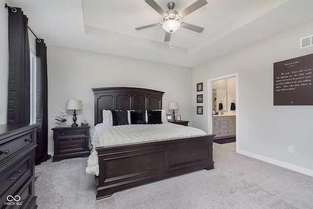 bedroom featuring a raised ceiling, a ceiling fan, ensuite bath, baseboards, and light colored carpet