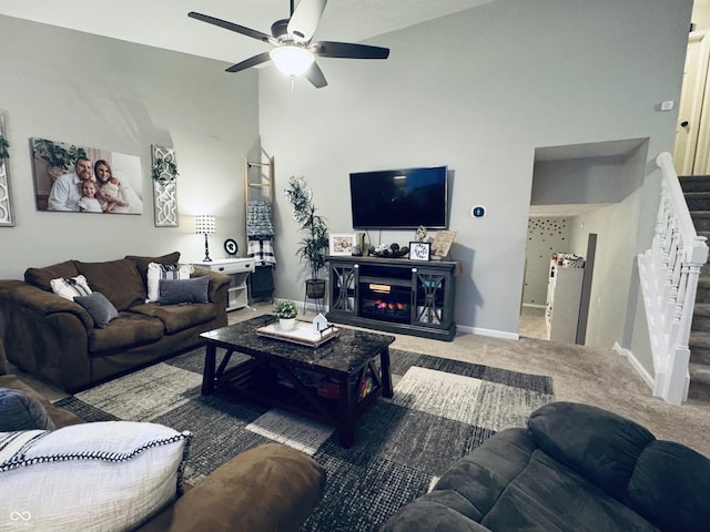 carpeted living room with baseboards, stairway, a high ceiling, a glass covered fireplace, and a ceiling fan