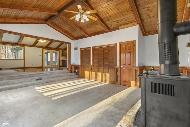 unfurnished living room featuring a wainscoted wall, a wood stove, carpet floors, wooden ceiling, and vaulted ceiling with beams