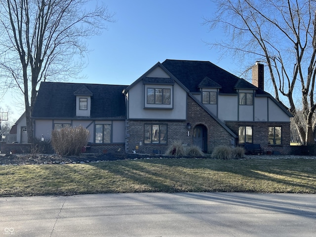 tudor-style house featuring stucco siding, a front yard, a shingled roof, brick siding, and a chimney