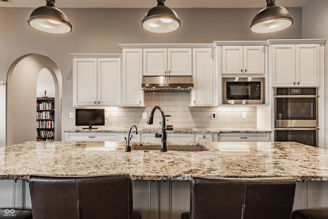 kitchen with stainless steel double oven, an island with sink, built in microwave, a sink, and under cabinet range hood