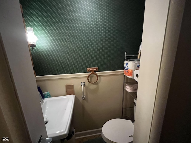 half bath with a sink, baseboards, toilet, and wallpapered walls