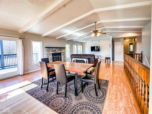 dining room with baseboards, light wood-type flooring, lofted ceiling with beams, a stone fireplace, and a textured ceiling