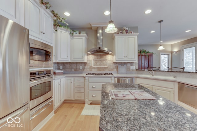 kitchen with stainless steel appliances, dark stone counters, light wood-style floors, wall chimney range hood, and decorative backsplash