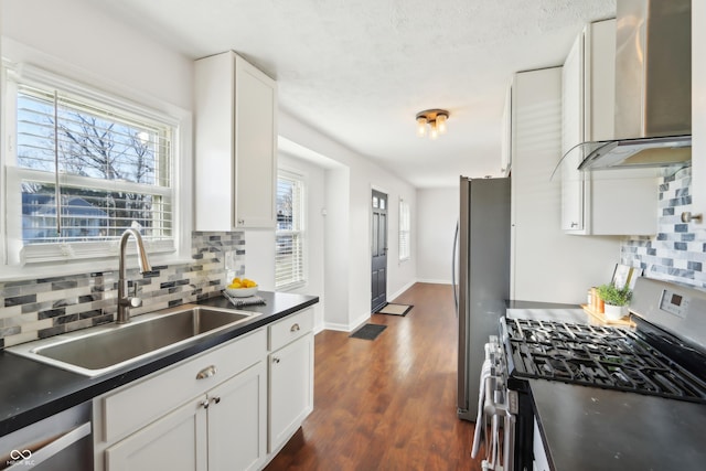 kitchen featuring a sink, stainless steel appliances, white cabinetry, dark countertops, and wall chimney range hood