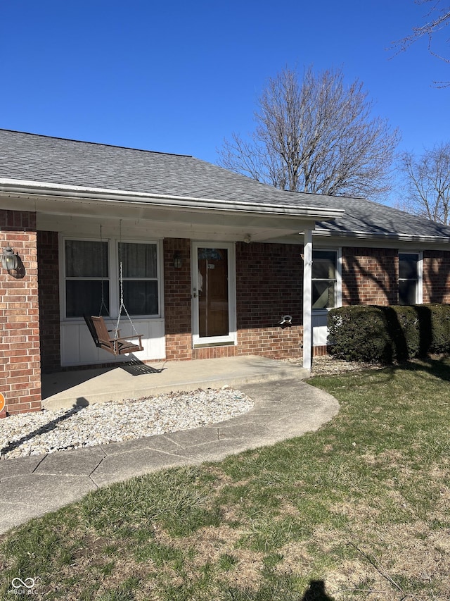 doorway to property featuring a yard, brick siding, and a shingled roof