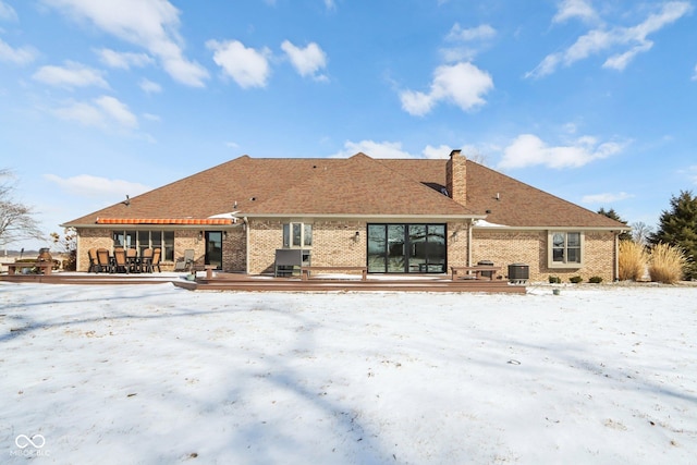 snow covered back of property with a deck, cooling unit, brick siding, and a chimney