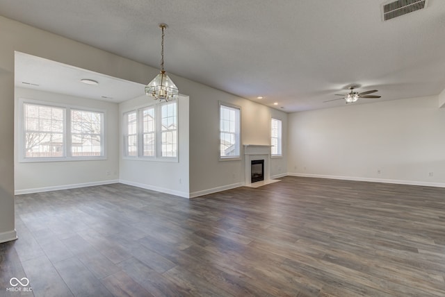 unfurnished living room featuring visible vents, baseboards, a fireplace with flush hearth, ceiling fan with notable chandelier, and dark wood-style floors