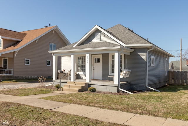 bungalow featuring covered porch, a front lawn, roof with shingles, and fence