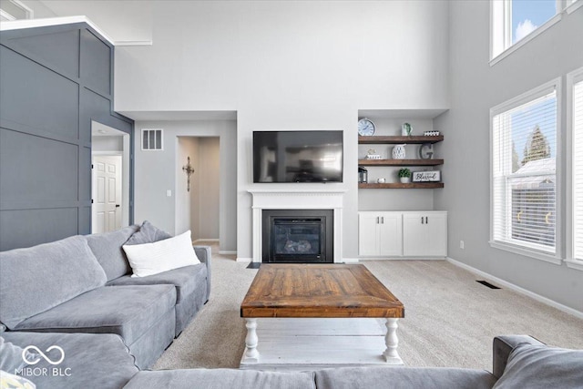 living room with visible vents, carpet flooring, a fireplace with flush hearth, and a towering ceiling