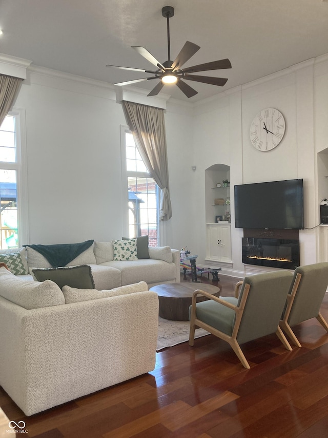 living room with built in shelves, dark wood-type flooring, ornamental molding, a ceiling fan, and a glass covered fireplace