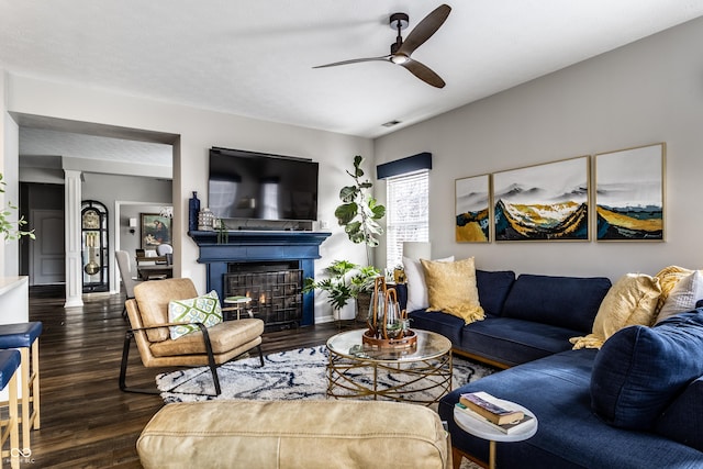 living room with dark wood finished floors, a glass covered fireplace, a ceiling fan, and ornate columns