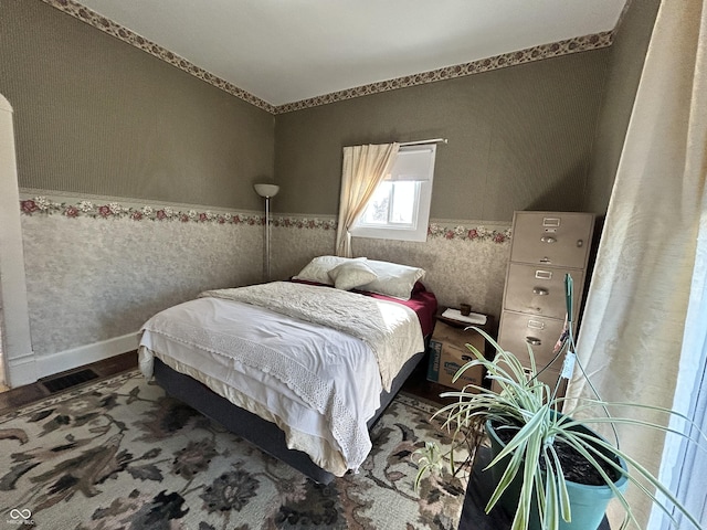 bedroom with wood finished floors, a wainscoted wall, baseboards, visible vents, and wallpapered walls