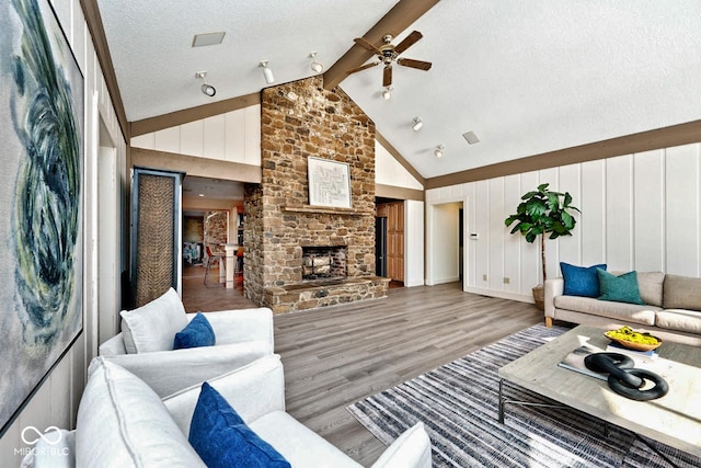 living area featuring beam ceiling, a textured ceiling, wood finished floors, a stone fireplace, and ceiling fan