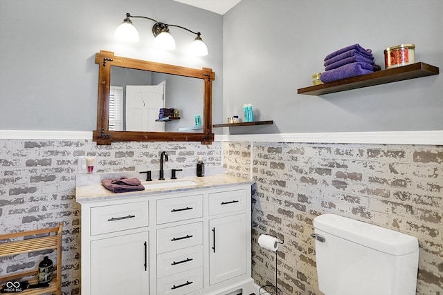 bathroom featuring wainscoting, toilet, brick wall, and vanity