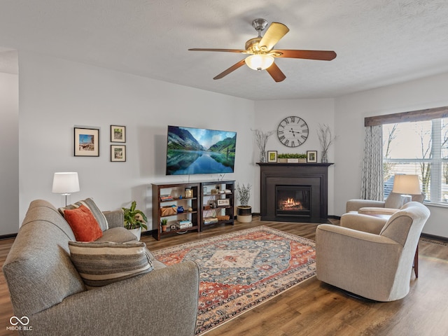 living area featuring ceiling fan, a textured ceiling, a warm lit fireplace, and wood finished floors