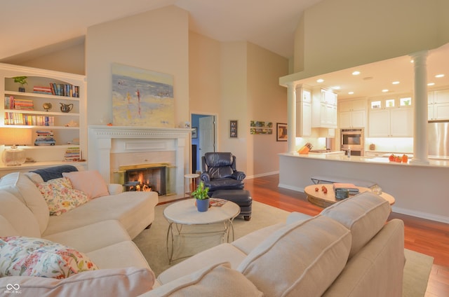 living area featuring high vaulted ceiling, ornate columns, baseboards, wood finished floors, and a glass covered fireplace