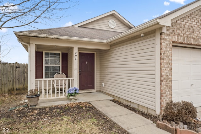 view of exterior entry featuring fence, roof with shingles, covered porch, an attached garage, and brick siding