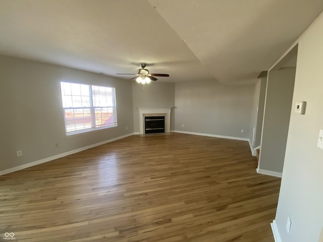 unfurnished living room featuring a ceiling fan, a fireplace with flush hearth, wood finished floors, and baseboards