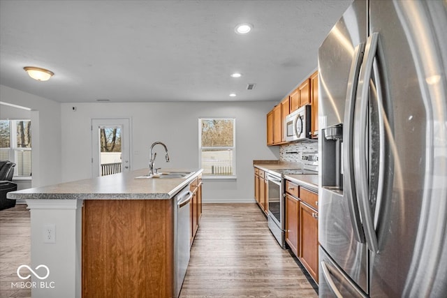 kitchen with a sink, light wood-type flooring, brown cabinetry, stainless steel appliances, and a kitchen island with sink