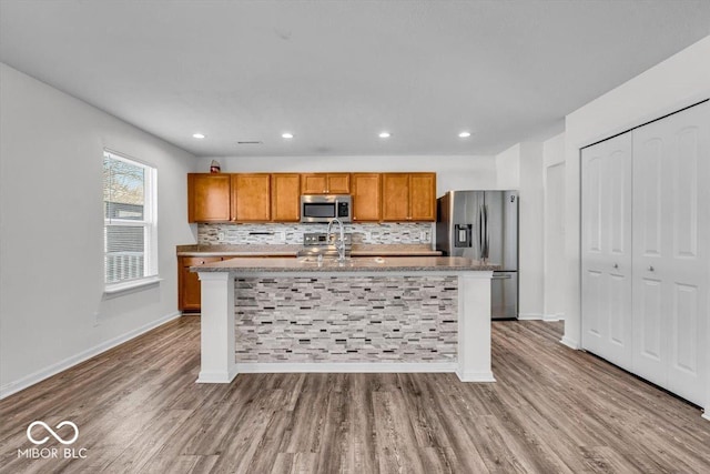 kitchen with wood finished floors, brown cabinetry, a kitchen island with sink, stainless steel appliances, and decorative backsplash