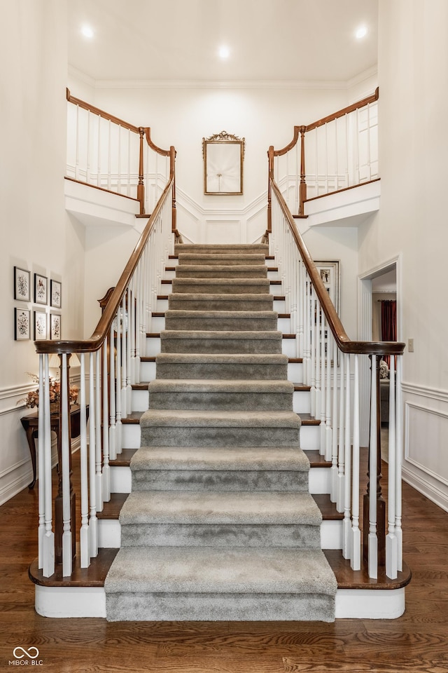 stairway featuring wood finished floors, a decorative wall, wainscoting, crown molding, and a towering ceiling