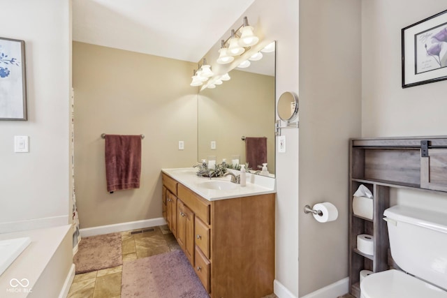 bathroom featuring toilet, a sink, stone tile floors, double vanity, and baseboards