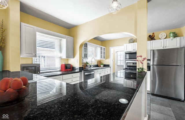 kitchen with dark stone countertops, arched walkways, white cabinets, stainless steel appliances, and a sink