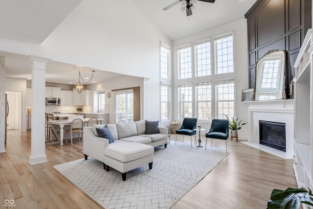 living area featuring a ceiling fan, high vaulted ceiling, light wood-style flooring, decorative columns, and a fireplace with flush hearth