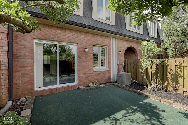 view of exterior entry featuring fence, brick siding, central AC, and roof with shingles