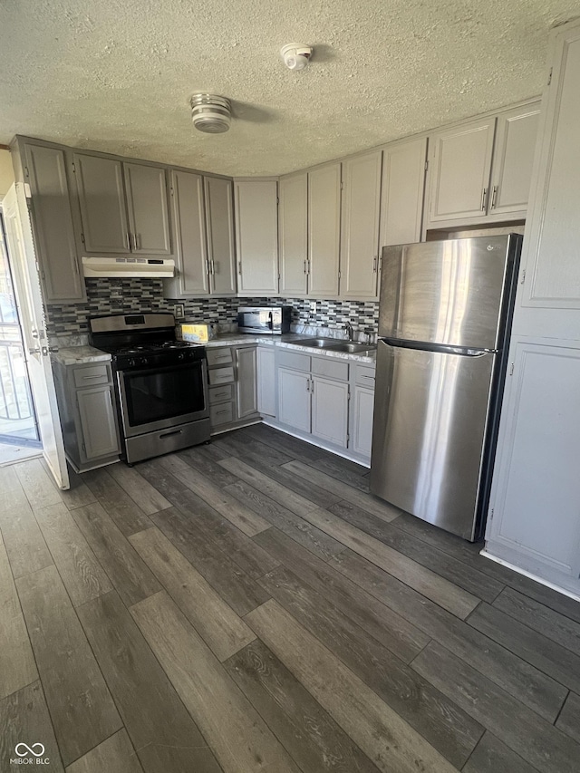 kitchen featuring dark wood finished floors, a sink, decorative backsplash, under cabinet range hood, and appliances with stainless steel finishes