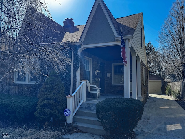 view of front of house with an outbuilding, covered porch, concrete driveway, roof with shingles, and a chimney