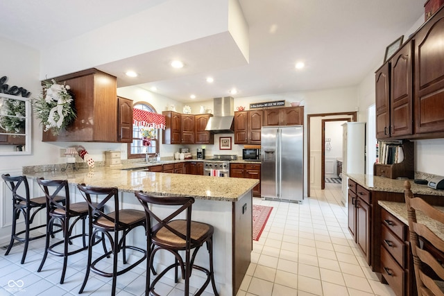 kitchen with a breakfast bar area, light stone counters, a peninsula, appliances with stainless steel finishes, and wall chimney range hood