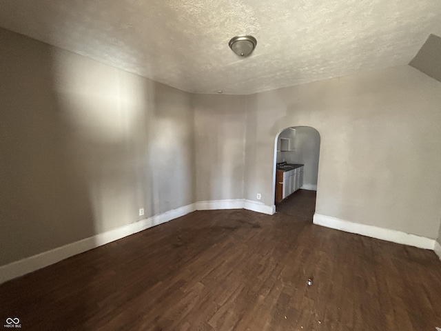 empty room featuring a textured ceiling, wood finished floors, arched walkways, baseboards, and vaulted ceiling