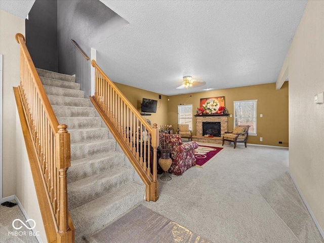 carpeted living area with baseboards, a textured ceiling, a stone fireplace, and stairs