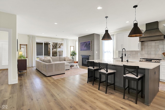 kitchen featuring premium range hood, light wood-style flooring, a kitchen island with sink, a sink, and light countertops