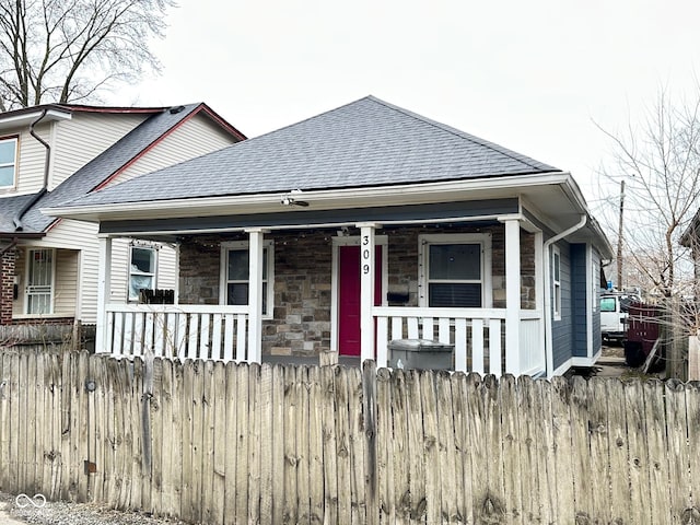 bungalow-style home with a porch, stone siding, a fenced front yard, and roof with shingles