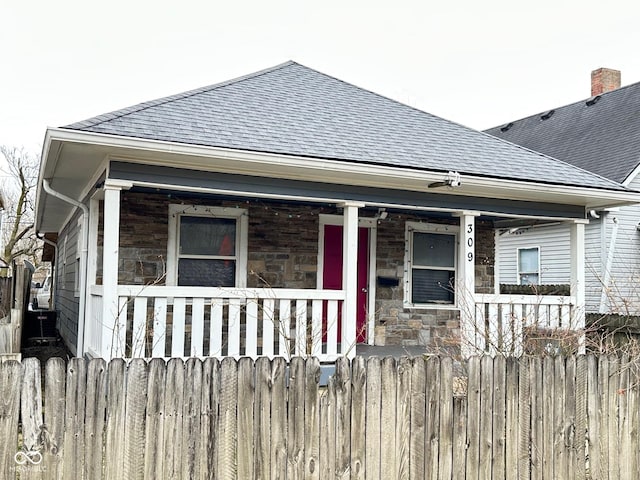 view of front facade featuring stone siding, fence, covered porch, and a shingled roof