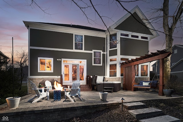 back of house at dusk with a hot tub, a fire pit, stone siding, a pergola, and a patio