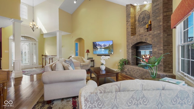 living room featuring a brick fireplace, decorative columns, wood finished floors, and high vaulted ceiling