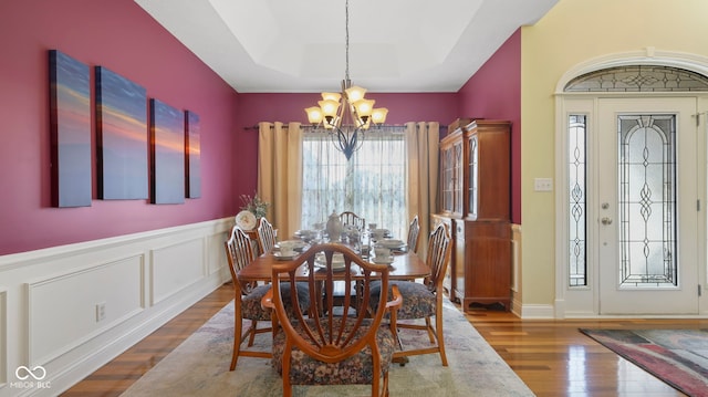 dining space with wood finished floors, wainscoting, a raised ceiling, a decorative wall, and a notable chandelier