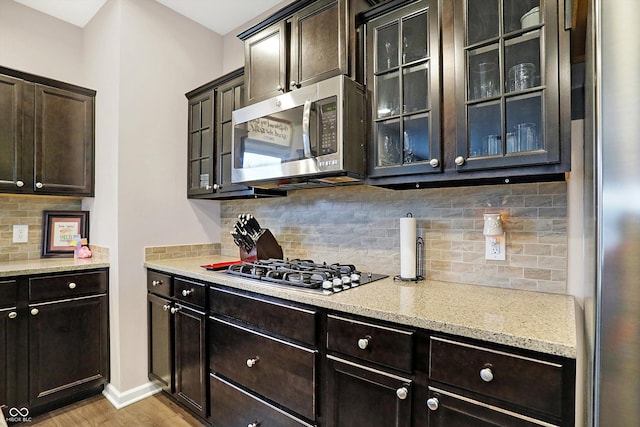 kitchen featuring light stone counters, backsplash, stainless steel appliances, light wood finished floors, and glass insert cabinets