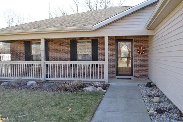 view of exterior entry featuring brick siding, covered porch, and a shingled roof