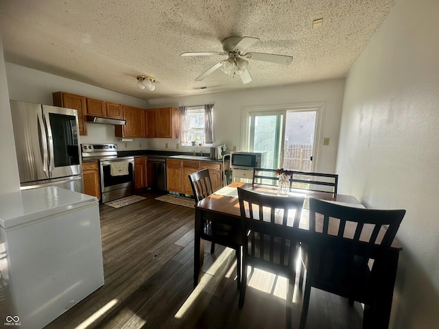 kitchen with a sink, under cabinet range hood, brown cabinets, stainless steel appliances, and dark wood-style flooring