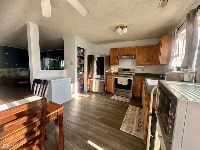 kitchen featuring brown cabinets, dark wood-type flooring, under cabinet range hood, a textured ceiling, and stainless steel appliances