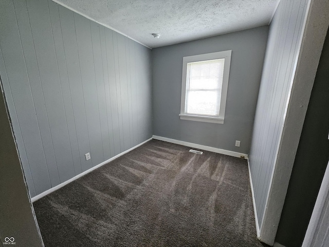 unfurnished room featuring visible vents, a textured ceiling, wooden walls, dark colored carpet, and baseboards